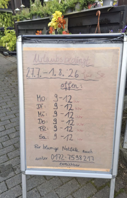 Handgeschriebenes Aufstellschild vor einem Blumengeschäft mit geänderten Öffnungszeiten: Urlaubsbedingt vom 27.7. bis 1.8.2026 von Montag bis Samstag jeweils von 9 bis 12 Uhr geöffnet. Für blumige Notfälle unter der Telefonnummer 0172-7598217 erreichbar.